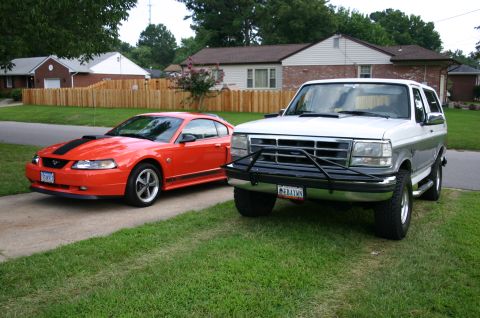 1994 Ford Bronco - An orange New Edge Ford Mustang coupe with a black hood stripe and hood scoop parked in a driveway beside a white full-size Ford Bronco fit…