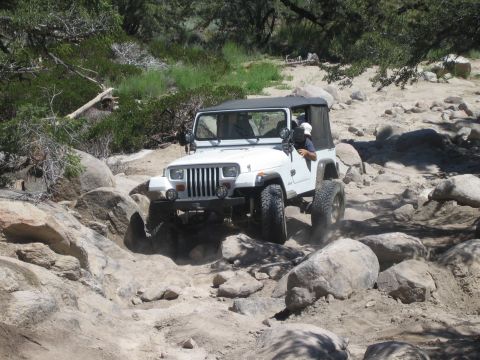 1990 Ford Bronco - white soft-top Jeep Wrangler with square headlights climbs boulders on a rocky desert trail.
