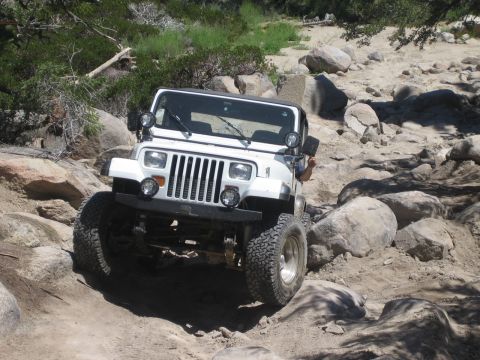 1990 Ford Bronco - white Jeep Wrangler with square headlights eases down a boulder-filled wash, a driver's arm out the window.