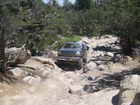 1990 Ford Bronco - dark green lifted SUV crawls along a rocky wash flanked by granite boulders and pines.