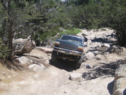1990 Ford Bronco - dark green lifted SUV with a front brush guard climbs over boulders on a sandy, tree-lined trail.