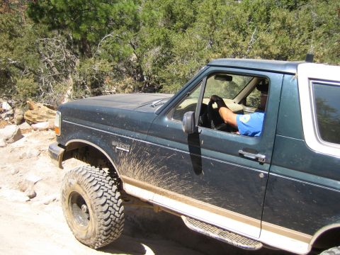 1990 Ford Bronco - lifted dark green Ford Bronco with mud on its flanks drives a dirt trail, the driver in a blue shirt visible inside.
