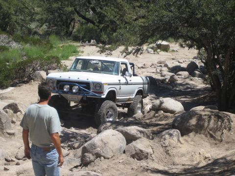 1990 Ford Bronco - white Ford pickup with FORD grille lettering sits among boulders in a dry sandy wash as a man stands watching at left.
