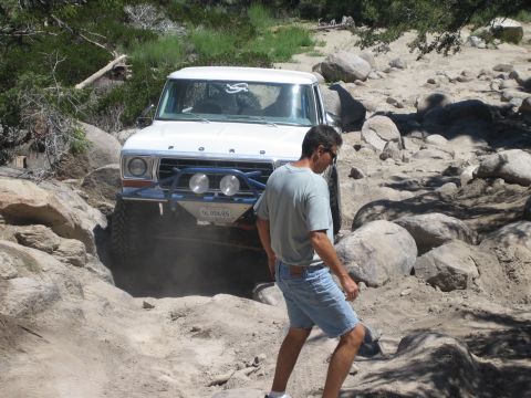 1990 Ford Bronco - white Ford truck with a blue tube bumper grips boulders on an off-road trail while a man in shorts walks alongside it.