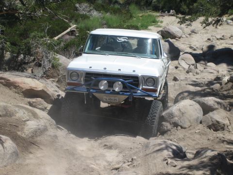 1990 Ford Bronco - white Ford truck with blue tube bumper and three round driving lights climbs over large boulders on a dusty trail.