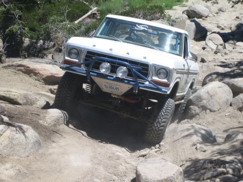 1990 Ford Bronco - white Ford truck with blue front bumper flexes its lifted suspension as it crawls over rocks, California plate visible.