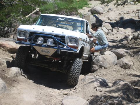 1990 Ford Bronco - man crouches beside the lifted front wheel of a white Ford truck as it crawls over boulders on a sandy trail.