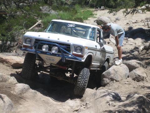 1990 Ford Bronco - man bends to inspect the front tire of a white Ford truck as it sits perched on boulders in a rocky wash.