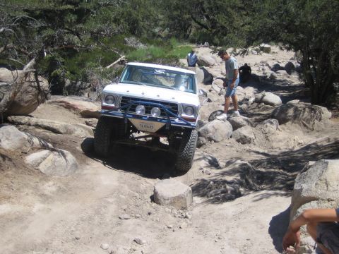 1990 Ford Bronco - white Ford truck noses down over a rock ledge on a dusty off-road trail as a man in shorts watches nearby.
