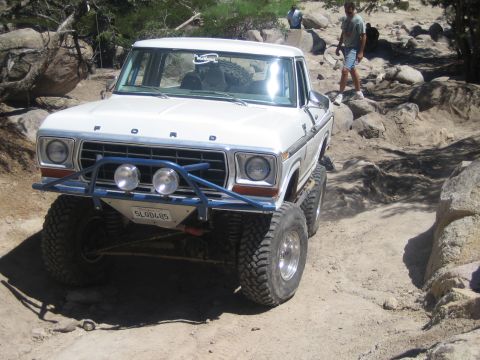 1990 Ford Bronco - white Ford truck with blue tube bumper and chrome wheels sits at an angle on a rocky trail with people in the background.