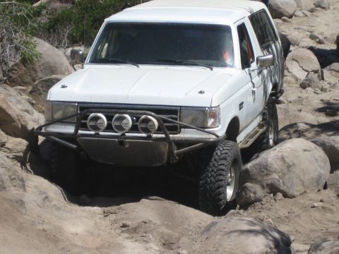 1990 Ford Bronco - white Ford Bronco with front light bar and lifted suspension articulates over boulders on a sandy off-road trail.