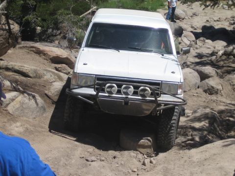 1990 Ford Bronco - white Ford SUV with a front light bar flexes its suspension as it drops into a rocky rut on an off-road trail.