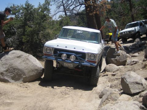 1990 Ford Bronco - white Ford truck with blue tube bumper squeezes between large rocks on a pine-shaded trail as people watch.