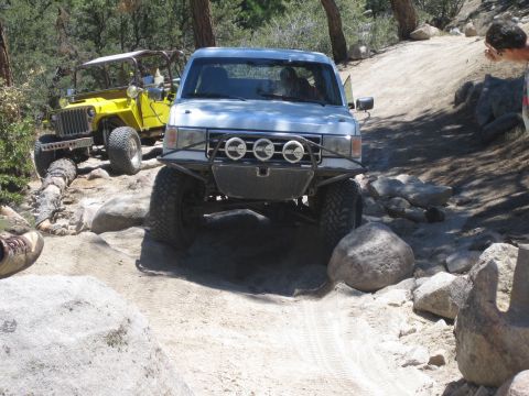 1990 Ford Bronco - silver-blue lifted truck with three driving lights crawls over rocks while a yellow flat-fender Jeep waits behind.