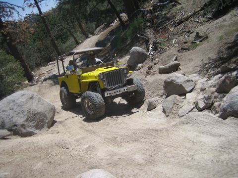 1990 Ford Bronco - yellow flat-fender Jeep with its headlights lit descends a steep dusty trail beside large boulders and pines.