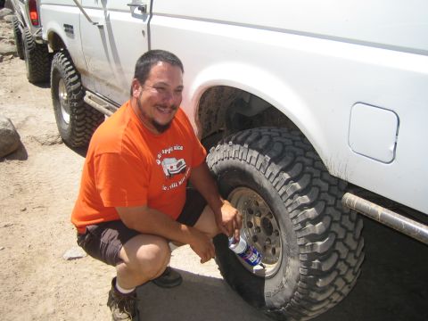 1990 Ford Bronco - smiling man in an orange graphic tee kneels beside the rear wheel of a white truck holding a can.