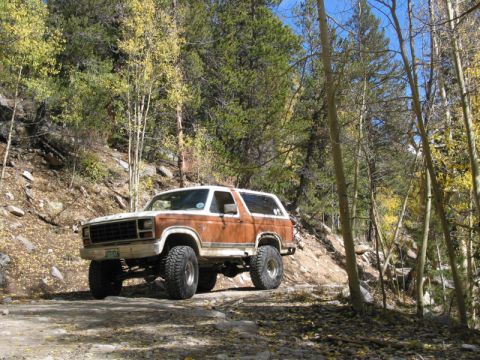 1981 Ford Bronco - A lifted Ford Bronco navigates a wooded dirt road during autumn.