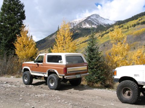 1981 Ford Bronco - A brown Ford Bronco is parked on a dirt road with autumn foliage and a snow-capped mountain in the distance.