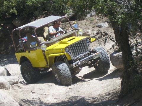 1990 Ford Bronco - man drives a yellow flat-fender Jeep with a rollbar and big tires over rocks on a dusty trail, license plate visible.