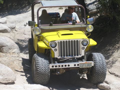 1990 Ford Bronco - yellow flat-fender Jeep with a vanity plate reading CUT LUUS climbs a rocky trail with its driver behind the wheel.