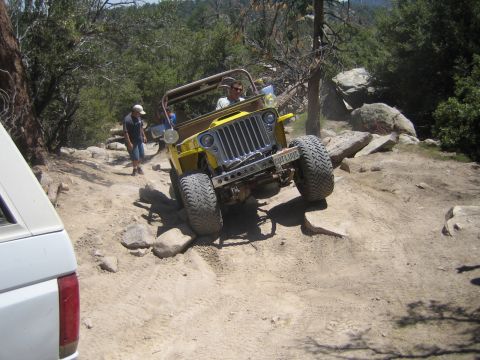 1990 Ford Bronco - yellow flat-fender Jeep crawls down a rocky trail as a person spots from the side and a white truck waits at left.