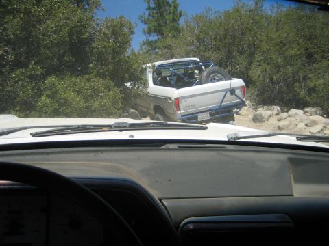 1990 Ford Bronco - Shot through a dusty windshield and wiper blades, a white pickup with an exposed roll bar and bed-mounted spare crawls a rocky desert trail.