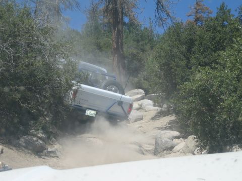1990 Ford Bronco - white Ford pickup with a blue tube rear bumper kicks up dust climbing a steep rocky obstacle among tall pines.