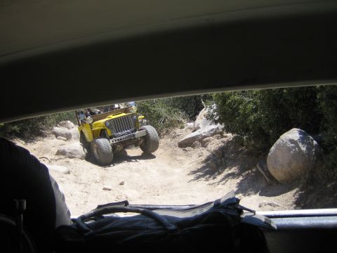 1990 Ford Bronco - Framed by a vehicle's roof and seat, a yellow flat-fender open-top jeep sits on a sandy boulder-strewn trail.