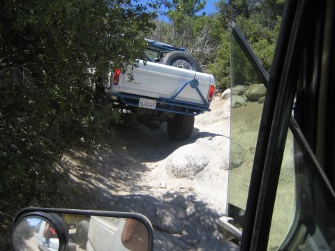 1990 Ford Bronco - Seen past an open door and side mirror, a lifted white Ford pickup with a blue tube bumper navigates a rocky ledge on a forest trail.
