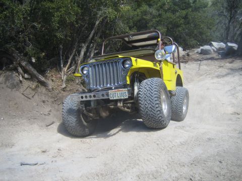 1990 Ford Bronco - bright yellow flat-fender open-top jeep with a roll bar, chrome wheels, and a personalized plate sits on a sandy trail.
