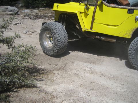 1990 Ford Bronco - close-up of a yellow open-top jeep's front fender, exposed exhaust, chrome wheel, and large off-road tire on rocky ground.