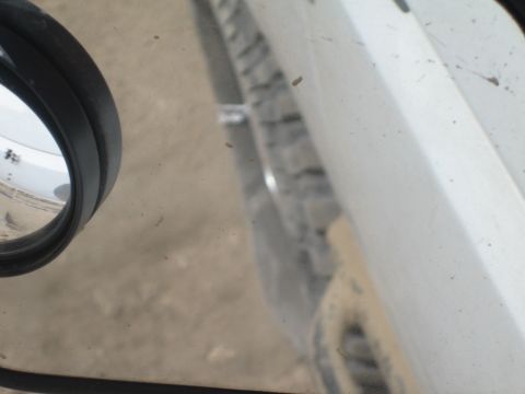 1990 Ford Bronco - blurry, tilted close-up showing a vehicle's round side mirror, white body panel, and tire above dusty ground.