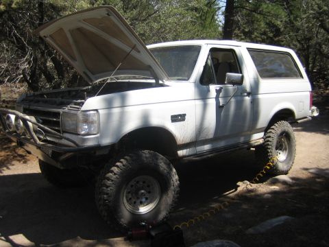 1990 Ford Bronco - white lifted Ford Bronco with its hood raised and a yellow tow strap attached to the front sits on a dirt clearing in the woods.