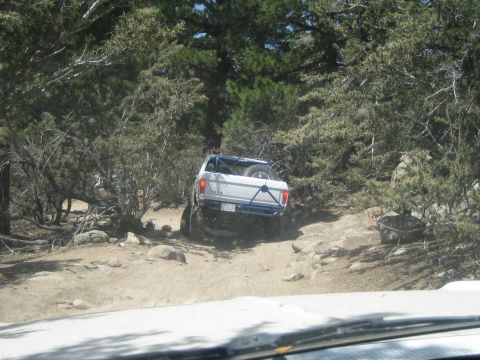 1990 Ford Bronco - white lifted pickup with a blue tube rear bumper crawls over rocks on a wooded trail, viewed over the photographer vehicle's hood.