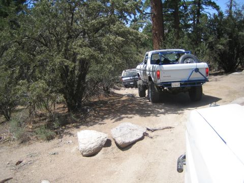 1990 Ford Bronco - white Ford pickup with a bed roll bar and spare tire travels a sandy trail past large boulders and brush in a pine forest.
