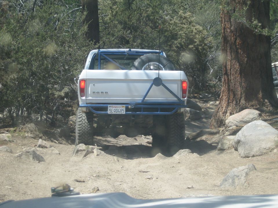 Rear of white Ford pickup with blue tube bumper climbing rocks in pine forest
