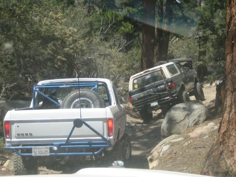 1990 Ford Bronco - white pickup with a blue tube bumper and a dark lifted SUV ahead of it crawl a rocky boulder section on a forested trail.