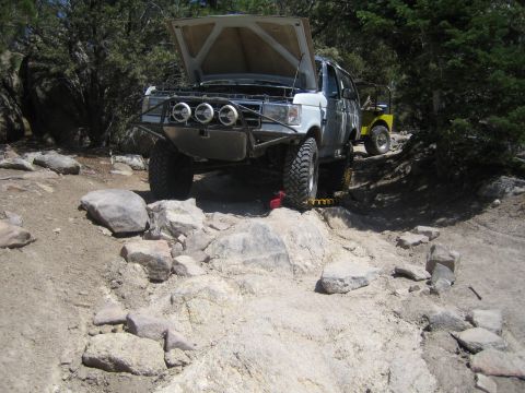 1990 Ford Bronco - lifted white Ford pickup with raised hood and brush guard sits on a boulder-strewn dirt trail while a yellow Jeep follows behind.