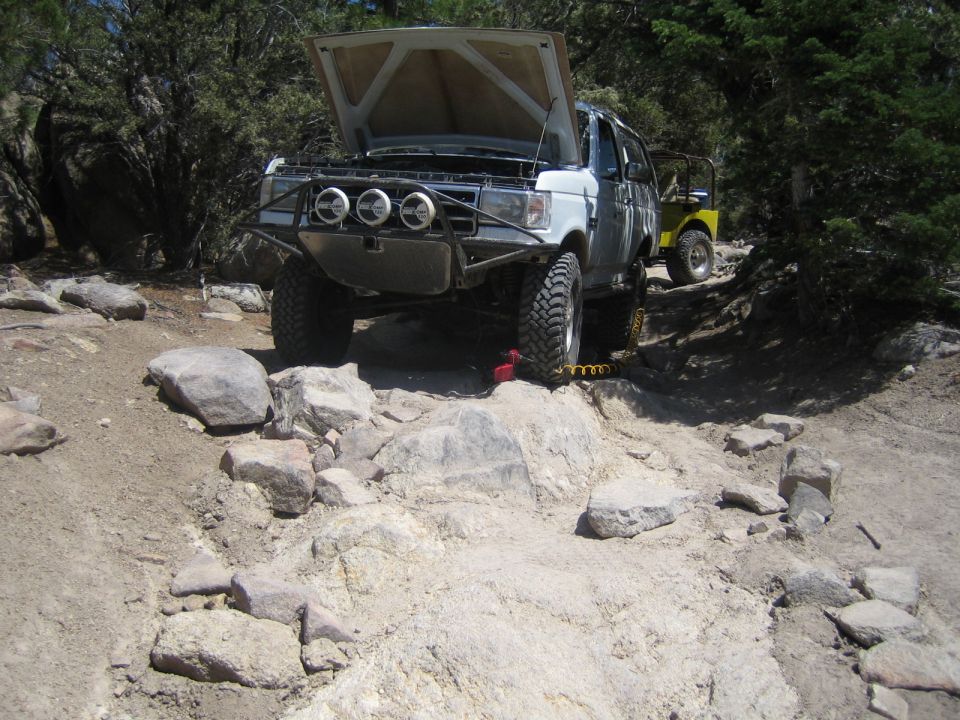 Lifted white Ford pickup with hood up and roof rack on a rocky off-road trail