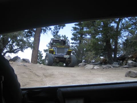 1990 Ford Bronco - yellow open-top Jeep with oversized tires crests a forested dirt trail, framed through the windshield of a following vehicle.