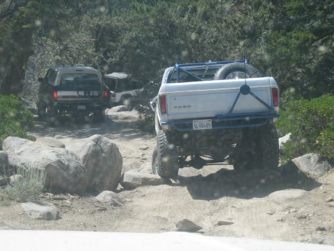 1990 Ford Bronco - lifted white Ford pickup with blue tube bumper and spare tire climbs rocks alongside dark SUVs on a dusty mountain trail.