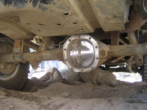 1990 Ford Bronco - dusty underside view of an off-road vehicle showing the chrome differential cover, axle and leaf-spring suspension over sandy ground.