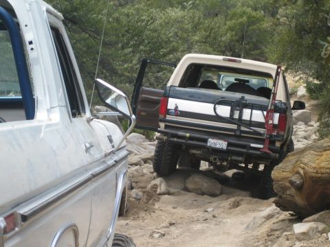 1990 Ford Bronco - two-tone tan-and-white Ford Bronco with an open door and rear tire carrier crawls over rocks next to a parked white Ford truck.