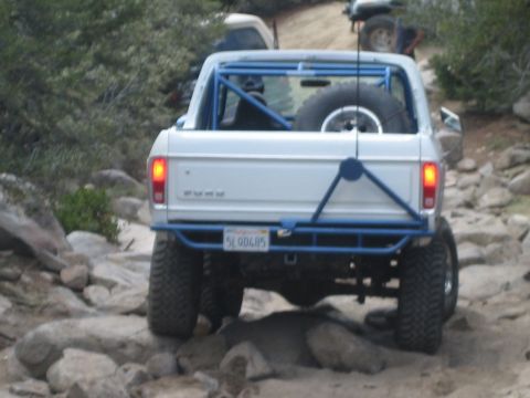 1990 Ford Bronco - lifted white Ford pickup with a blue roll cage, spare tire and glowing tail lights climbs a steep rocky obstacle at dusk.