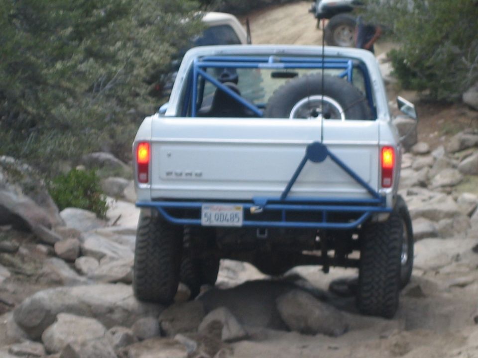 Rear of a lifted white truck with blue roll cage climbing a rocky hill