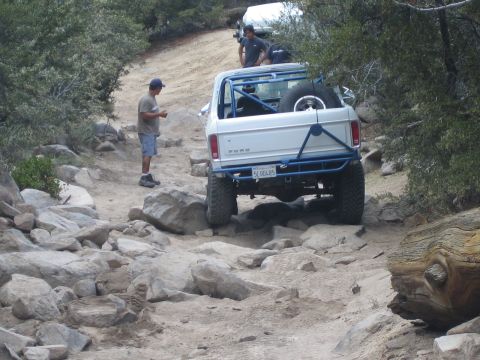 1990 Ford Bronco - Two spotters stand on a dusty trail watching a lifted white Ford pickup with a blue roll cage work through a rocky section.