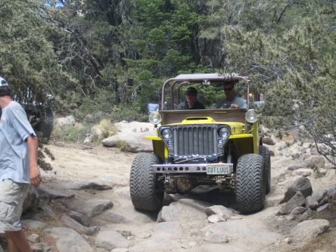 1990 Ford Bronco - yellow vintage flat-fender Jeep with a roll cage and CUTLUUS plate carries two occupants over rocks on a forested dirt trail.