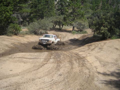 1990 Ford Bronco - white pickup truck splashes through a muddy water-filled rut on a sandy forested off-road trail, spraying mud from its tires.