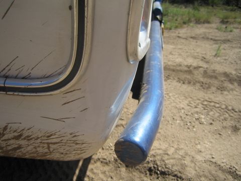 1990 Ford Bronco - Close-up of a heavily scratched white vehicle rear corner panel and the capped end of a blue steel tube bumper over dirt.
