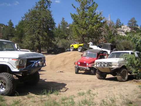 1990 Ford Bronco - group of off-road vehicles, including a white Ford truck, red Jeep and yellow Jeep, gather on a dirt clearing among pine trees.